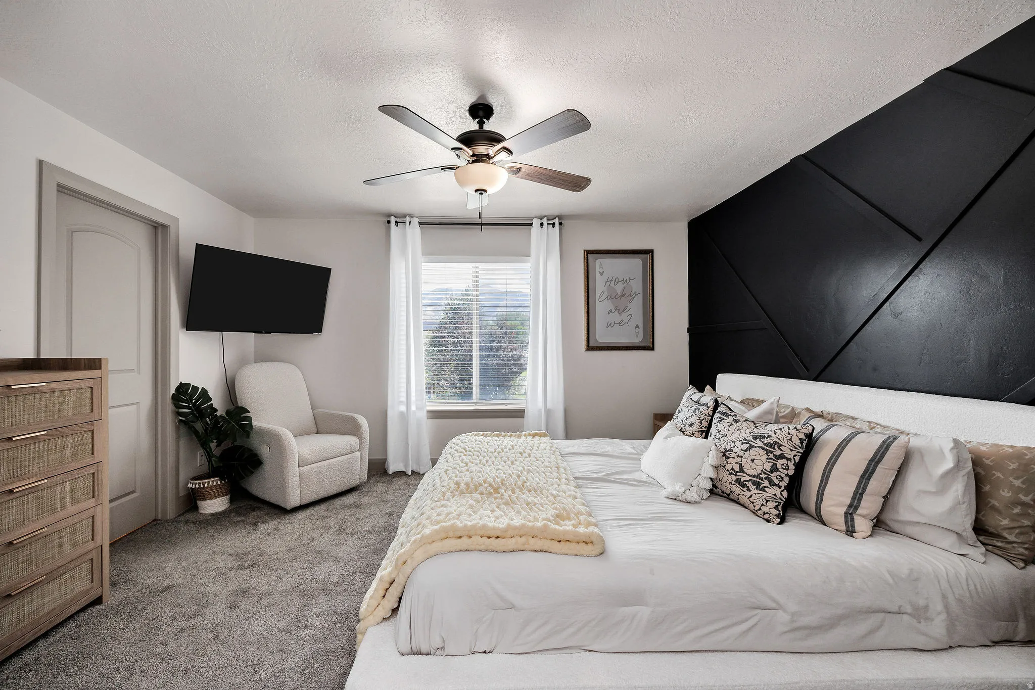 Textured accent wall, ceiling fan with integrated lighting, window with blinds, wall-mounted television, and neutral carpeting