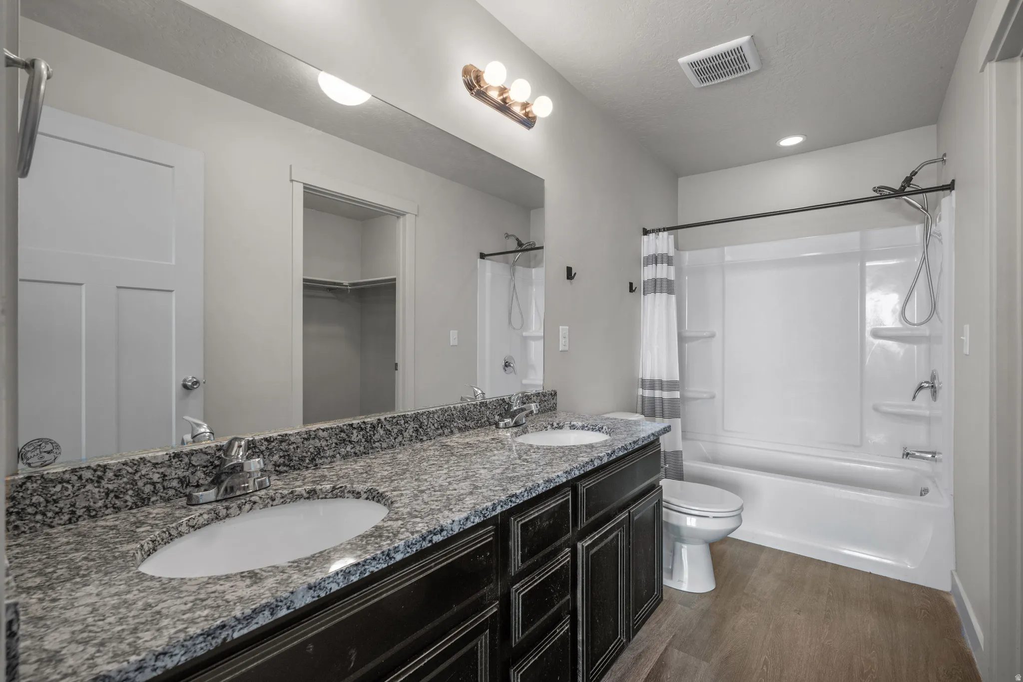 Double vanity with speckled countertop and under-mount sinks, wood-finish flooring, and a white shower-over-tub combination