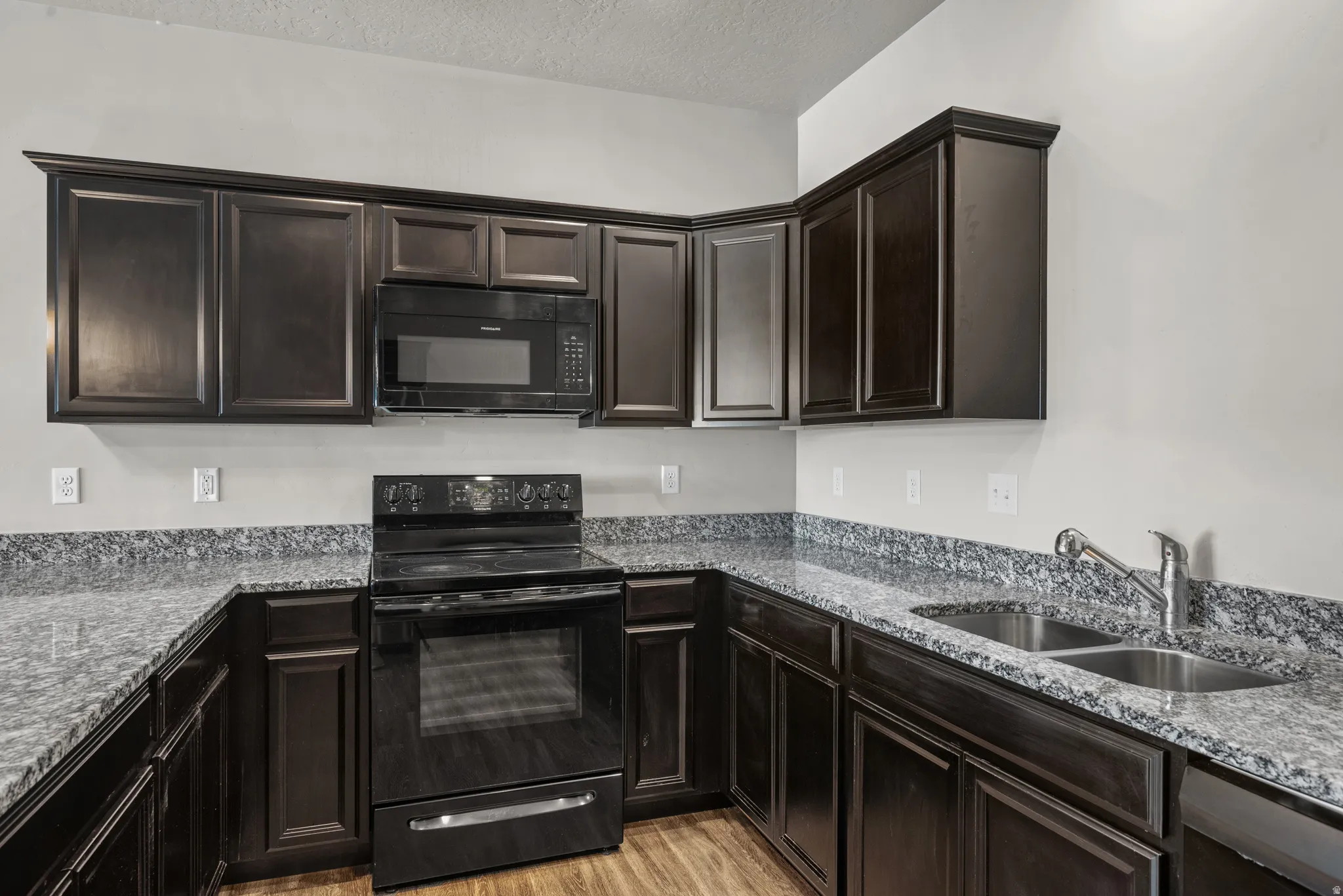 Kitchen featuring dark wood-finish cabinetry, speckled countertops, black electric range, black microwave, and a double basin stainless steel sink