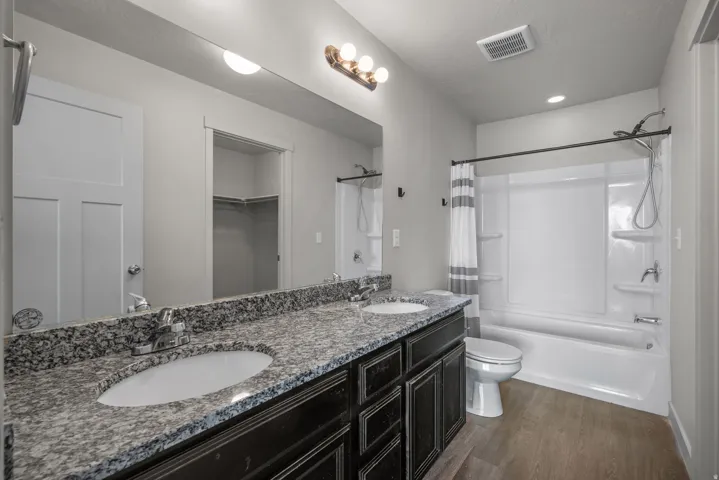 Double vanity with speckled countertop and under-mount sinks, wood-finish flooring, and a white shower-over-tub combination
