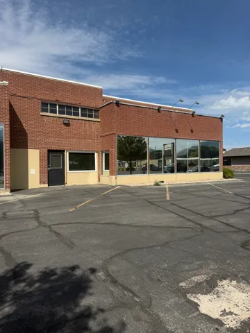 Brick exterior with multiple large storefront windows, secondary entry with black door, and expansive asphalt parking area