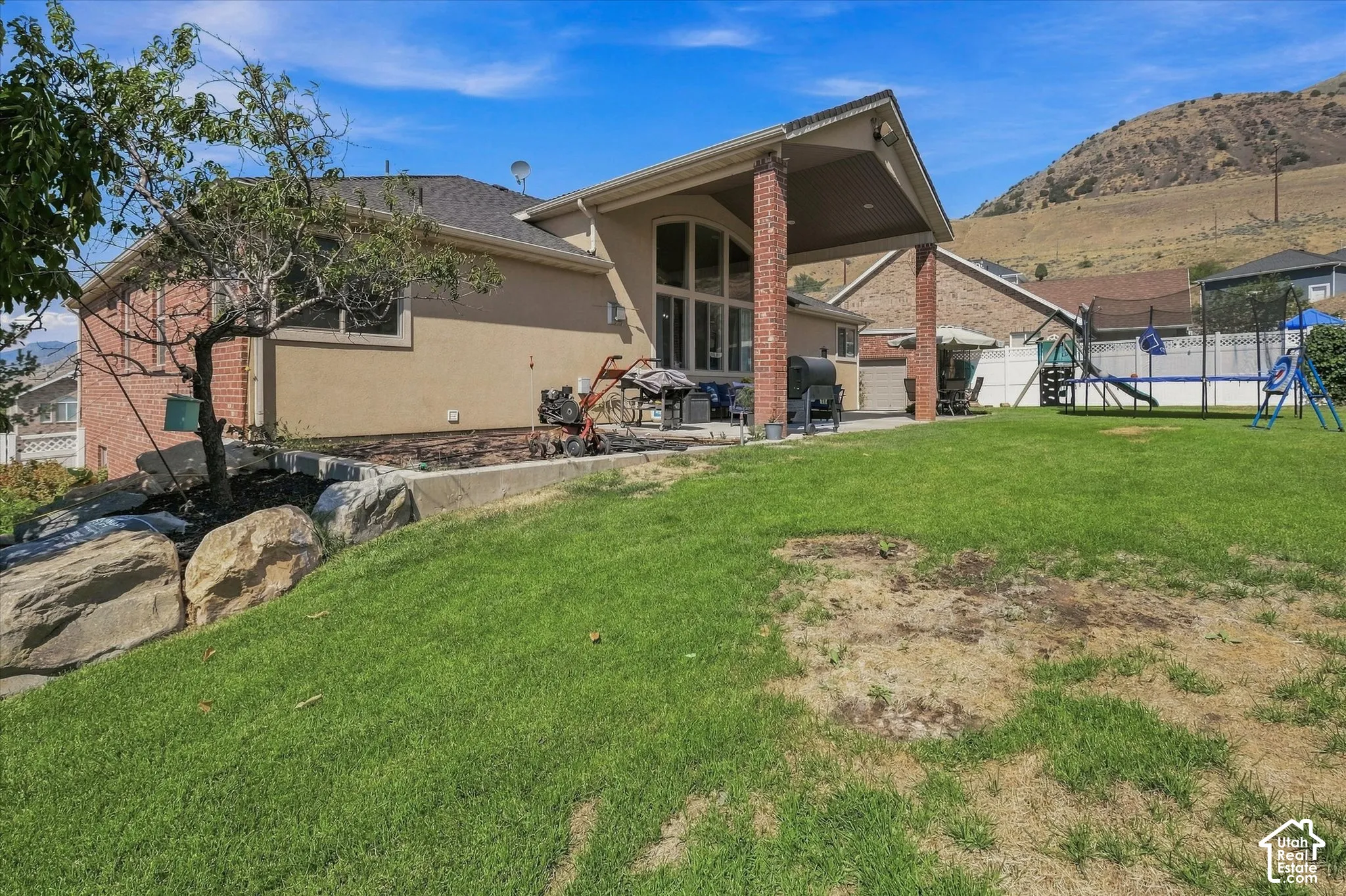 Expansive backyard featuring a covered patio with brick columns, a terraced landscape bed with large boulders, and a sloped grass area