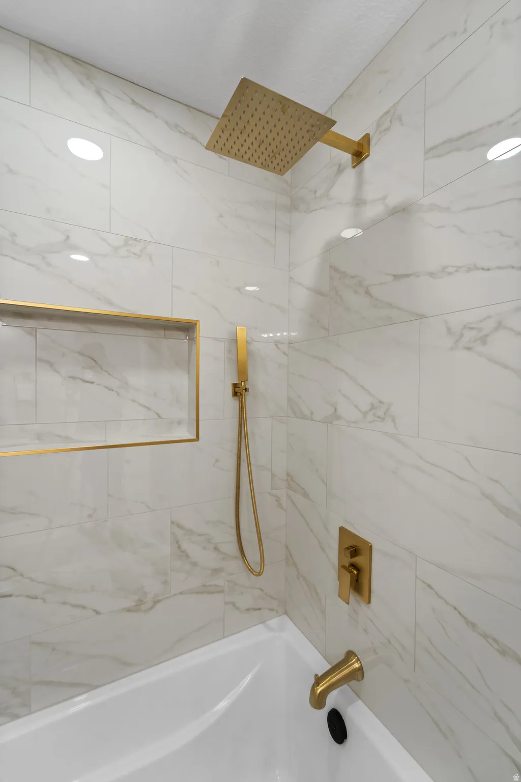 Bathtub with gold-finish fixtures, white marble-look wall tiling, recessed shower niche with gold-finish trim, and a rain showerhead