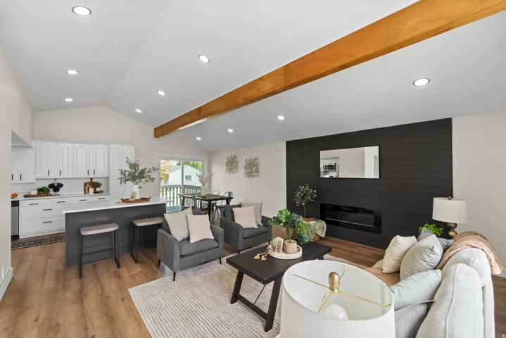 Vaulted ceiling with an exposed wood beam, contemporary electric fireplace with shiplap surround, wood-finish flooring, white cabinetry, and a kitchen island