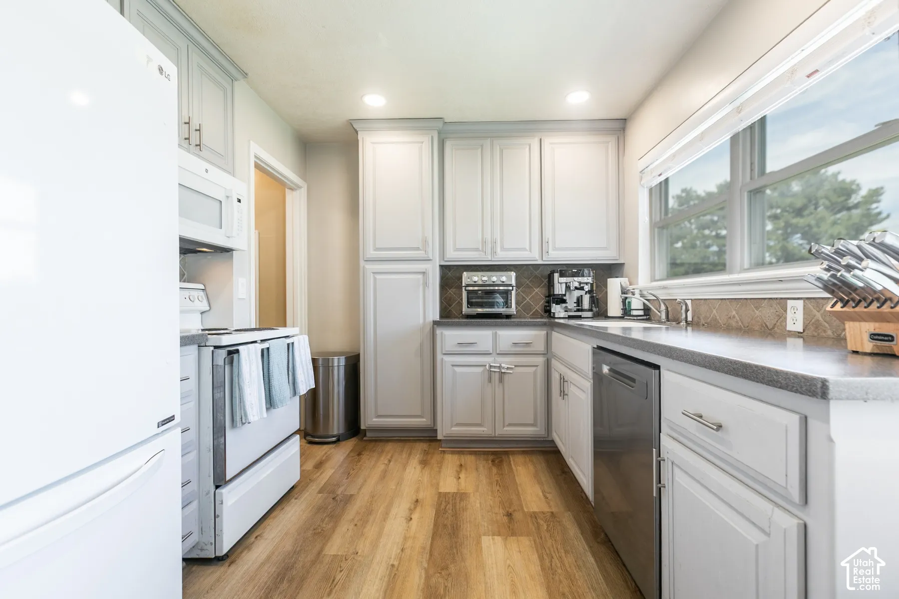 White cabinetry with brushed nickel hardware, wood-finish flooring, dark countertops, tiled backsplash, and recessed lighting