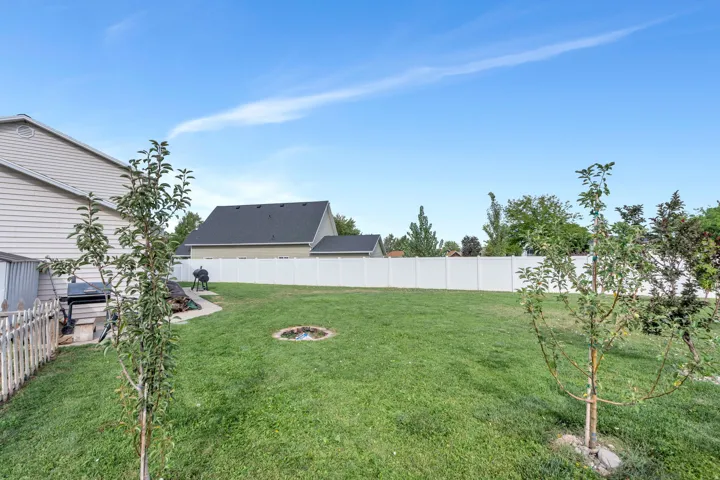 Expansive backyard featuring a manicured lawn, mature landscaping with newly planted trees, and a fire pit, backing to a white privacy fence