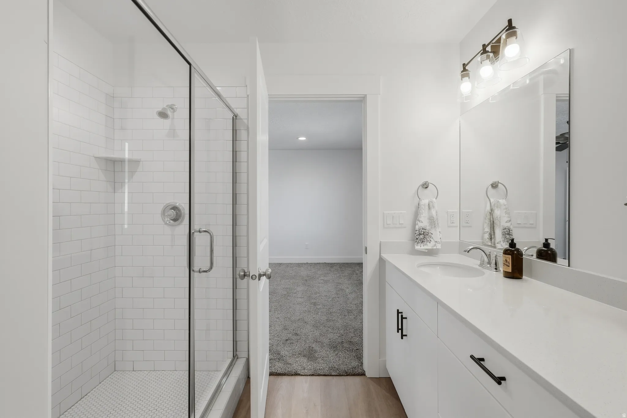 Walk-in shower with subway tile surround and glass enclosure, single vanity with integrated sink and chrome faucet, full-width mirrored wall, wood-finish flooring, and overhead vanity lighting