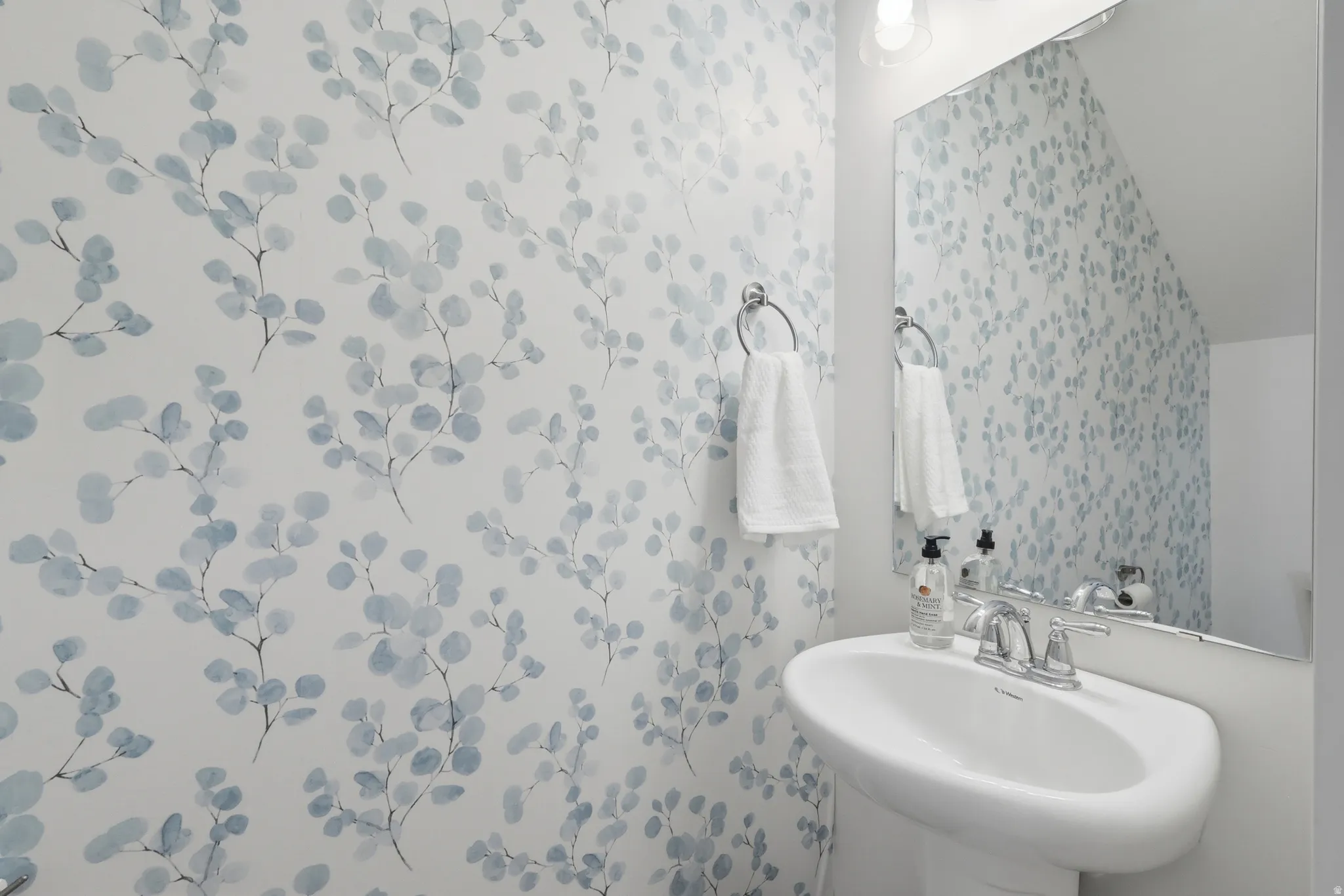 Pedestal sink with chrome fixtures and an oval mirror, complemented by botanical-patterned wallpaper