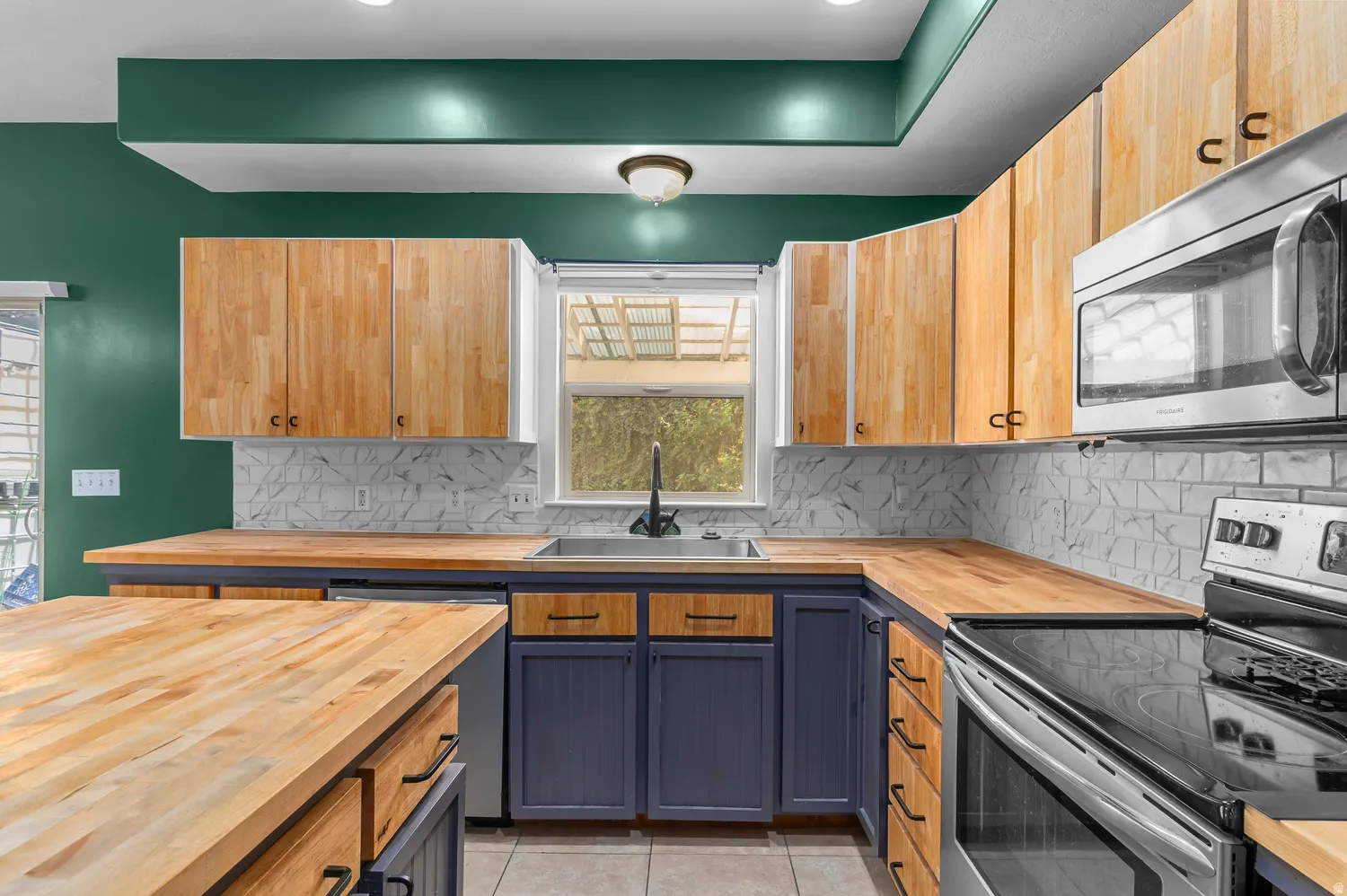 Two-tone cabinetry with wood-finish upper cabinets and blue lower cabinets, wood-finish countertops, geometric tile backsplash, and stainless steel appliances