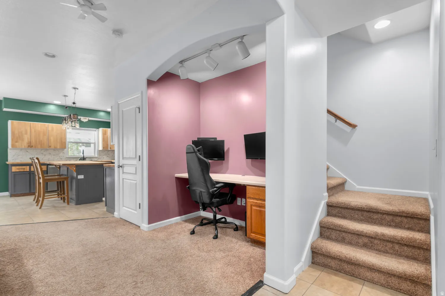 Carpeted living area with tile flooring entry, built-in desk with track lighting, and a staircase with a wooden handrail