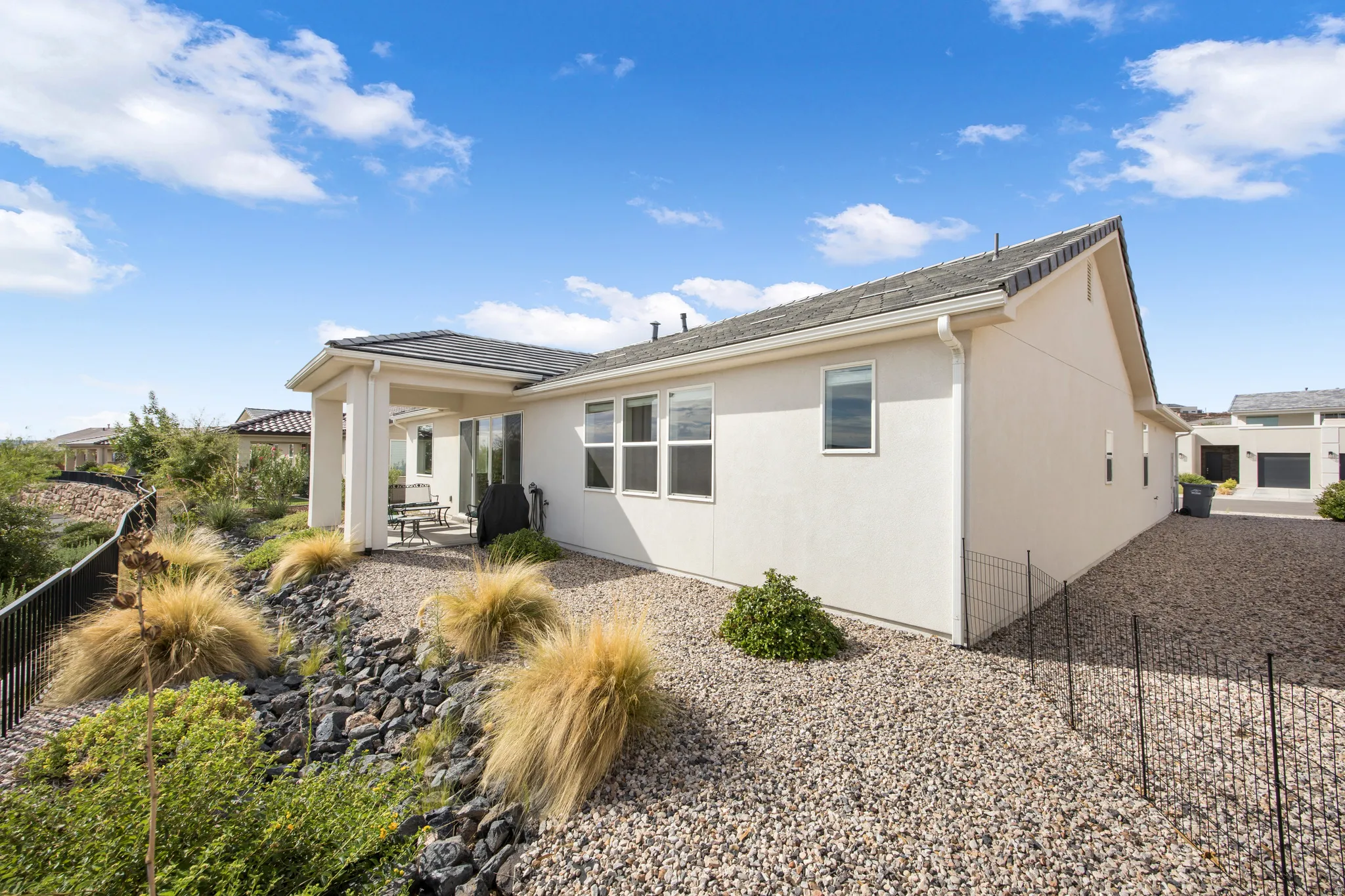 Exterior stucco finish, covered patio with support columns, multiple windows, gravel landscaping, and a low-maintenance sloped yard with natural rock features