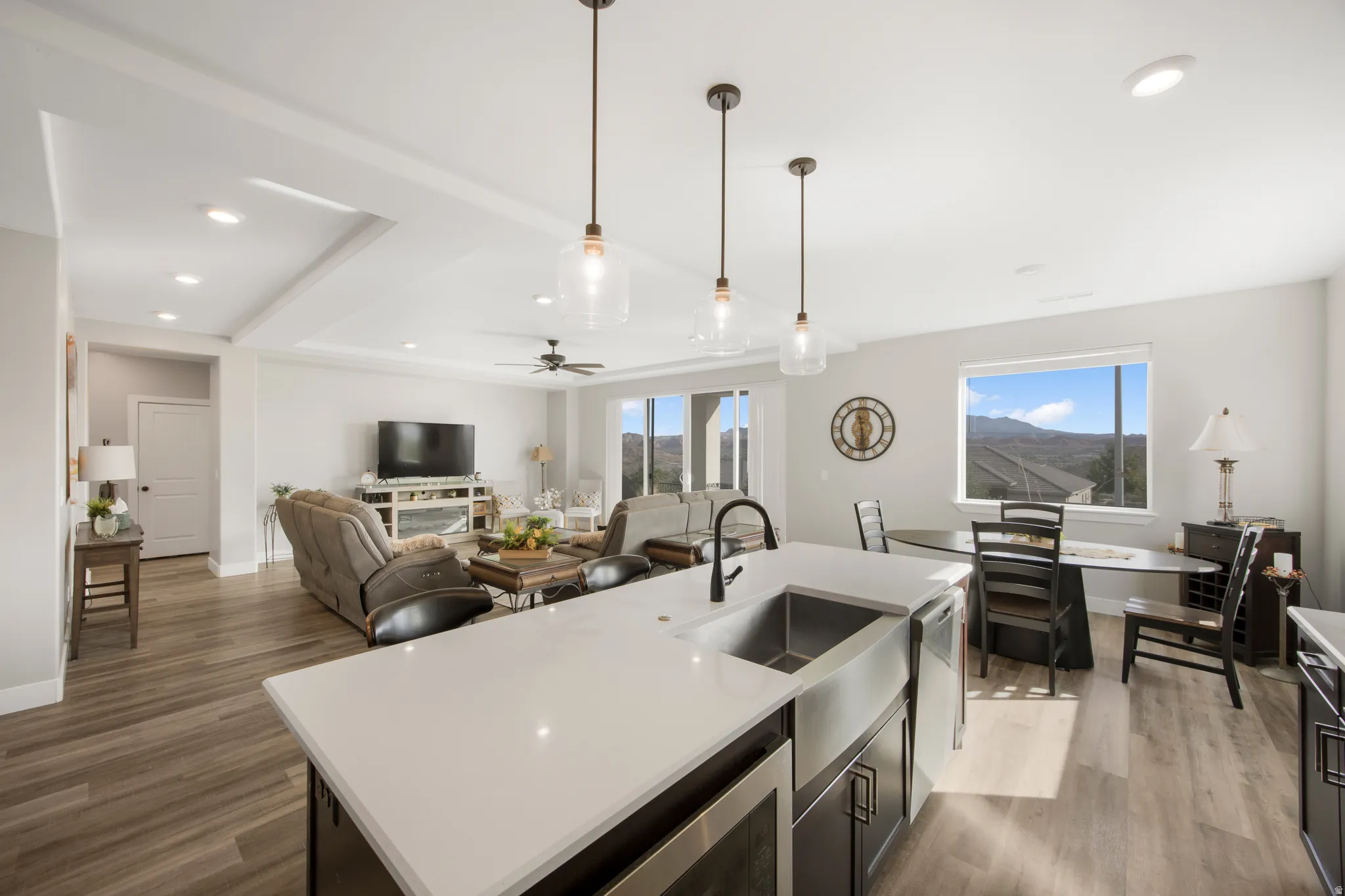 Open-concept living space with wood-finish flooring, a kitchen island with a farmhouse sink, pendant lighting, and a prominent window