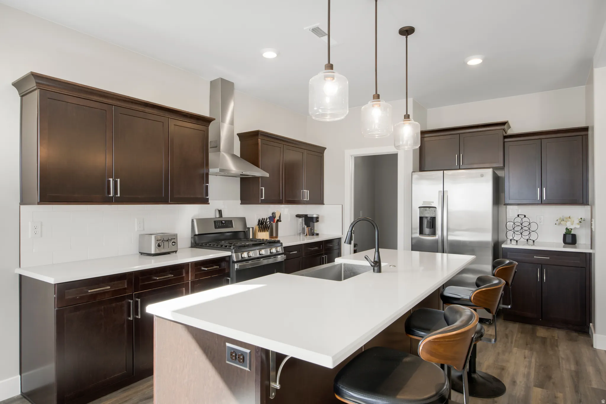 Kitchen featuring dark wood-finish cabinetry, stainless steel appliances, a white tile backsplash, and a central island with a sink and pendant lighting