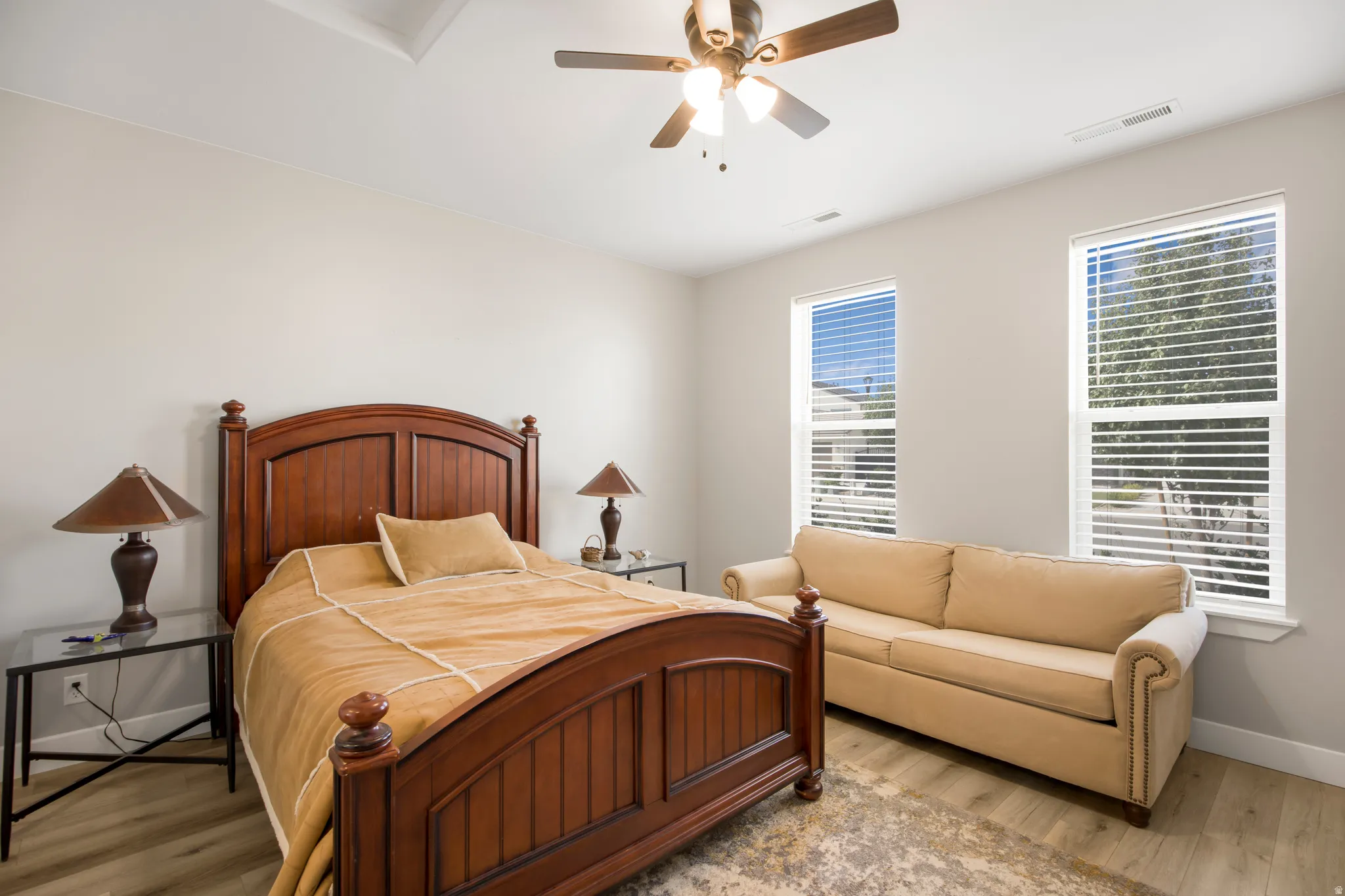 Wood-finish flooring, two large windows with blinds, a ceiling fan with integrated lighting, and light-colored wall paint