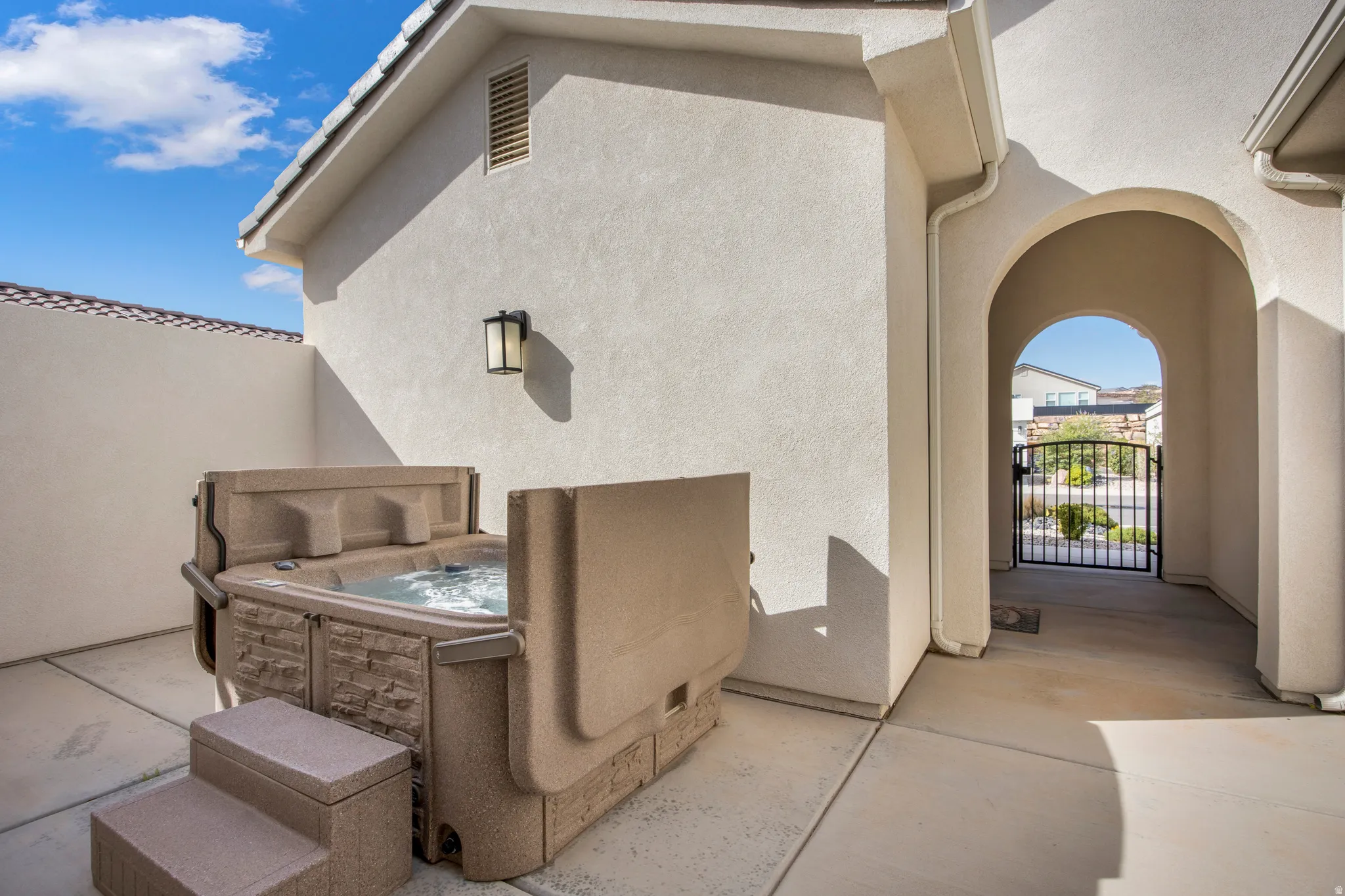 Private enclosed courtyard featuring a hot tub, concrete patio, stucco walls, and an arched entryway leading to a wrought iron gate