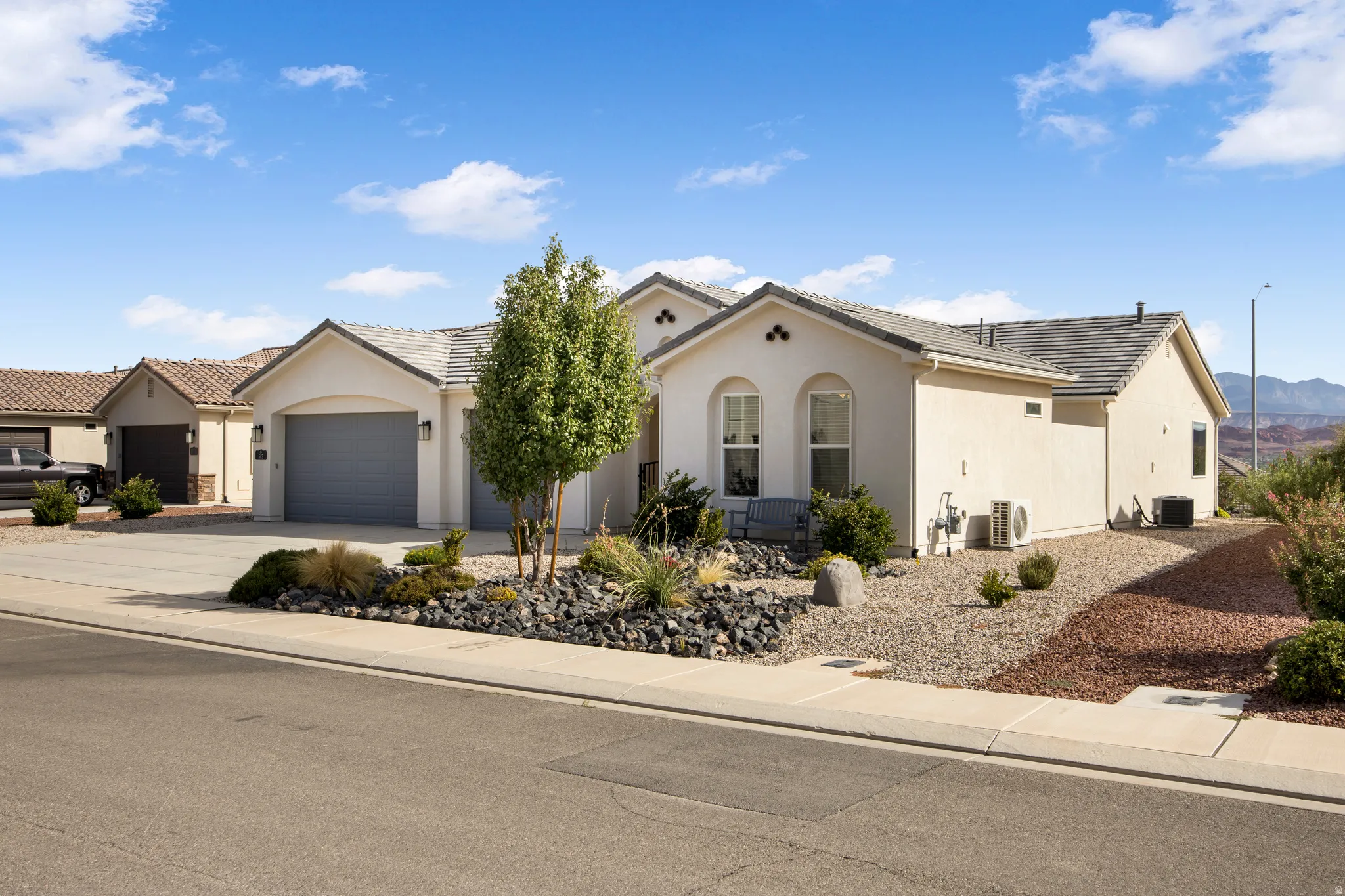 Elegant single-story residence featuring a neutral stucco exterior with arched architectural details, a three-car garage, and professional landscaping with desert-adapted plantings and decorative rock hardscaping