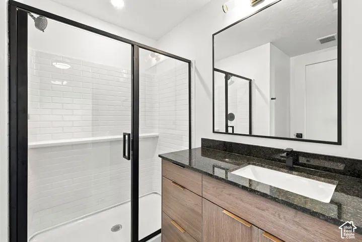 Bathroom vanity with wood-finish cabinetry, dark stone countertop, rectangular undermount sink, and matte black hardware, featuring a glass-enclosed shower with white subway-style tile and a black-framed mirror