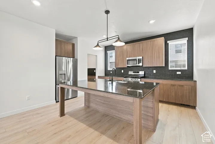 Kitchen featuring flat-panel wood-finish cabinetry, a large central island with a stone countertop, a contrasting dark brick backsplash, stainless steel appliances, and wood-finish flooring