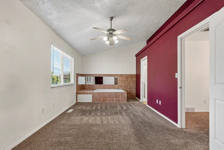 Vaulted ceiling with ceiling fan, recessed soaking tub with tiled surround, multiple mirrors, and a window with blinds