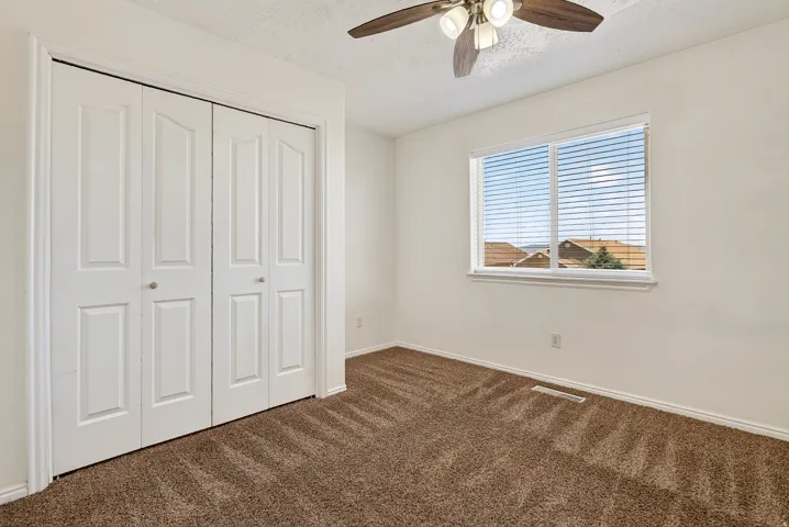 White bi-fold closet doors, a ceiling fan with light fixture, an expansive window with blinds, and brown carpet flooring