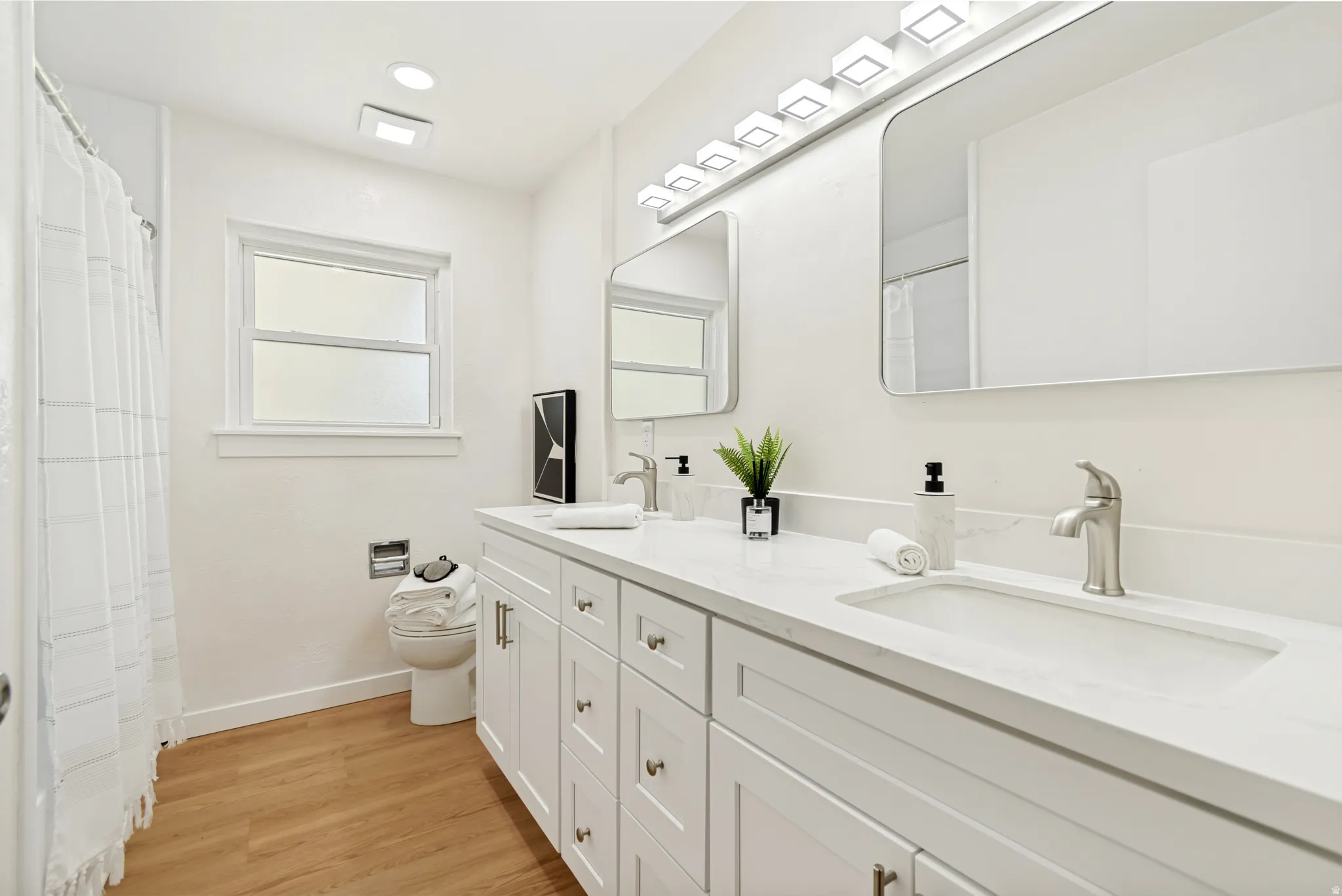 Double vanity with integrated sinks, white cabinetry, undermount lighting, wood-finish flooring, and a window