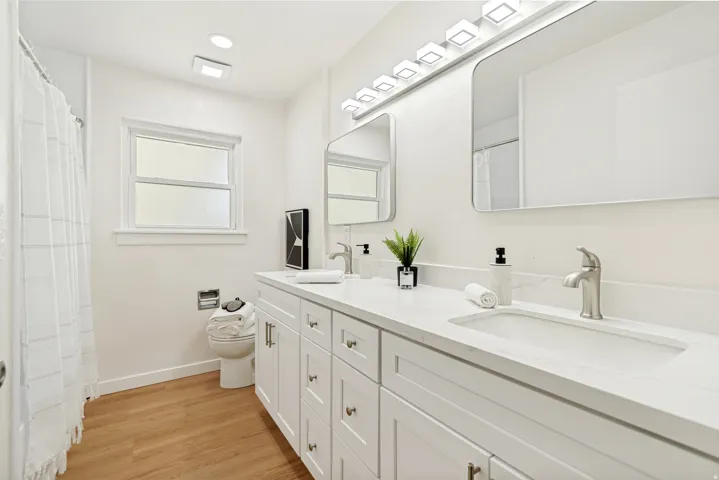 Double vanity with integrated sinks, white cabinetry, undermount lighting, wood-finish flooring, and a window
