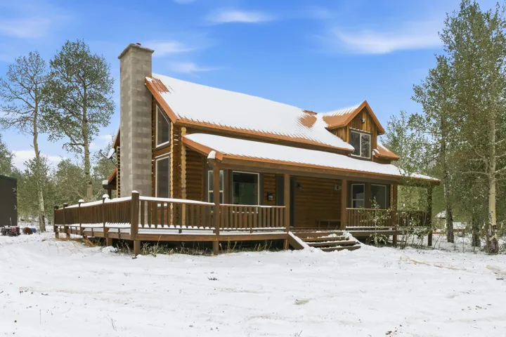 Log cabin exterior featuring a prominent stone chimney, a covered front porch, a wood deck, and a dormer window