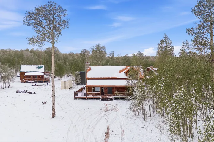 Two distinct structures with log-sided exteriors, covered front porches, and snow-dusted roofs, surrounded by mature trees