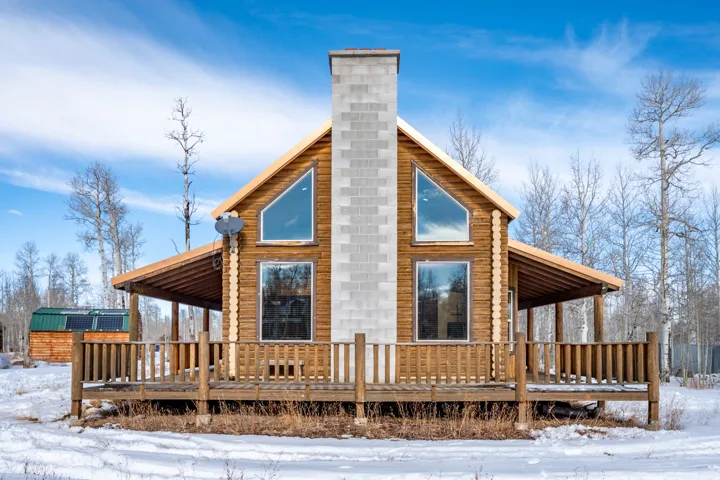 Log cabin exterior with a full-width covered porch, exposed wood beams, a stone-finish chimney, and prominent triangular and rectangular windows