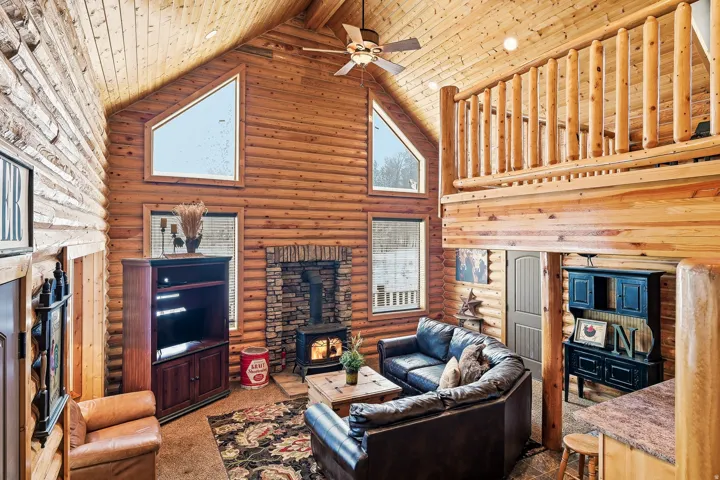 Vaulted ceiling with exposed wood beams, full-height stone fireplace with wood stove, wood-finish flooring, and a loft with wood railing