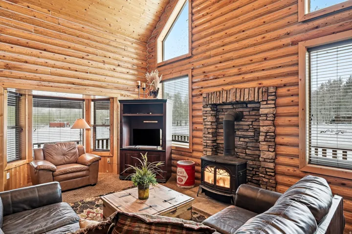 Vaulted wood-paneled ceiling, stone-clad fireplace with wood-burning stove, large picture windows, wood-paneled walls, and built-in media cabinet