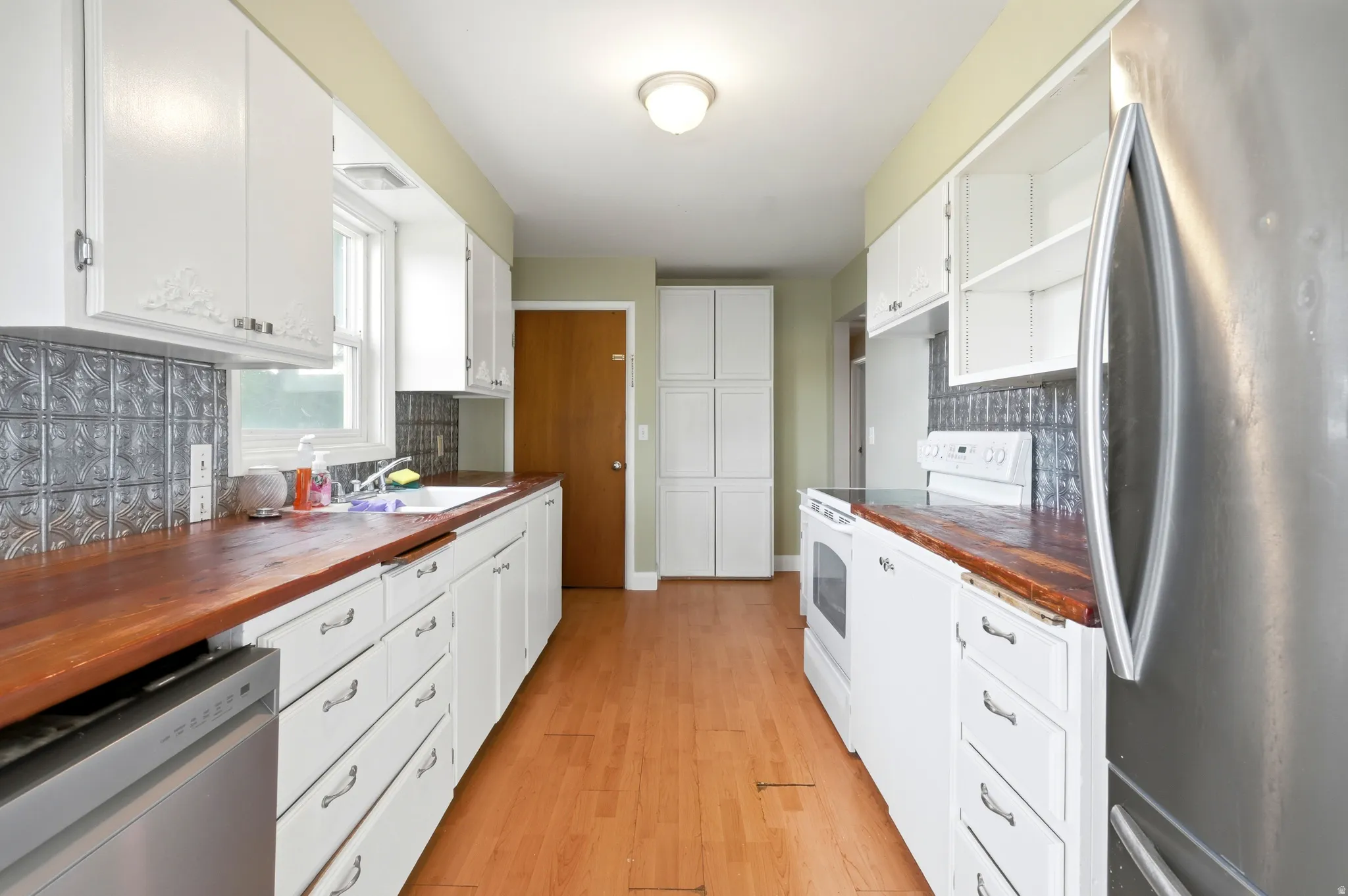 Galley kitchen featuring wood-finish countertops, white cabinetry, a tin-panel backsplash, hardwood-style flooring, and a stainless steel refrigerator