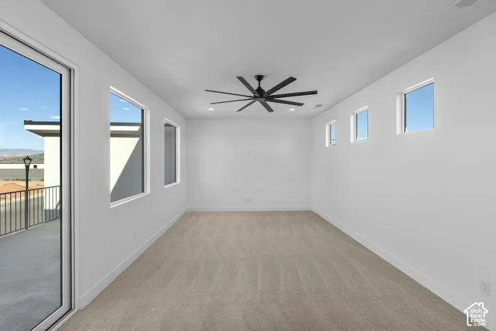 Spacious room featuring neutral carpeting, a modern ceiling fan, multiple windows, and a large glass door leading to a balcony with black metal railing