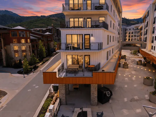 Modern multi-story building featuring balconies with glass railings, a ground-level patio, and a mountain backdrop