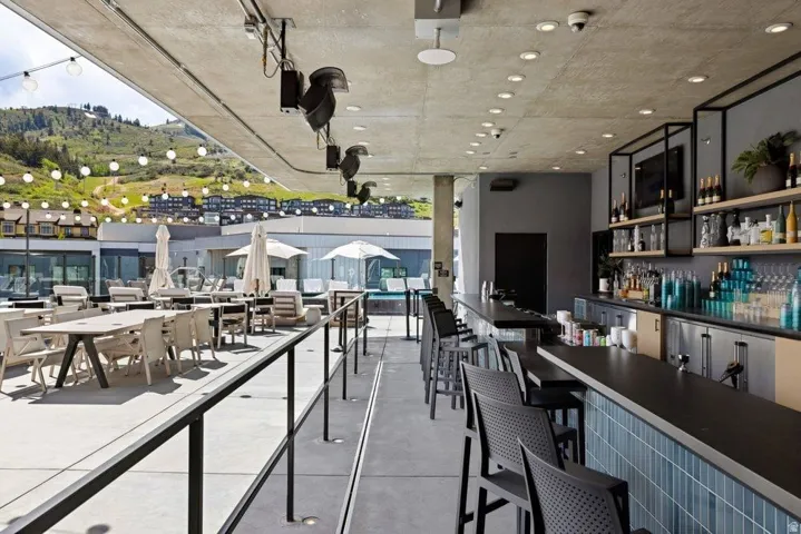 Modern rooftop bar and lounge featuring floor-to-ceiling glass railings, a full-service bar with tiered shelving and beverage display, concrete ceiling with recessed lighting, and expansive outdoor seating areas with panoramic vistas of the surrounding hi