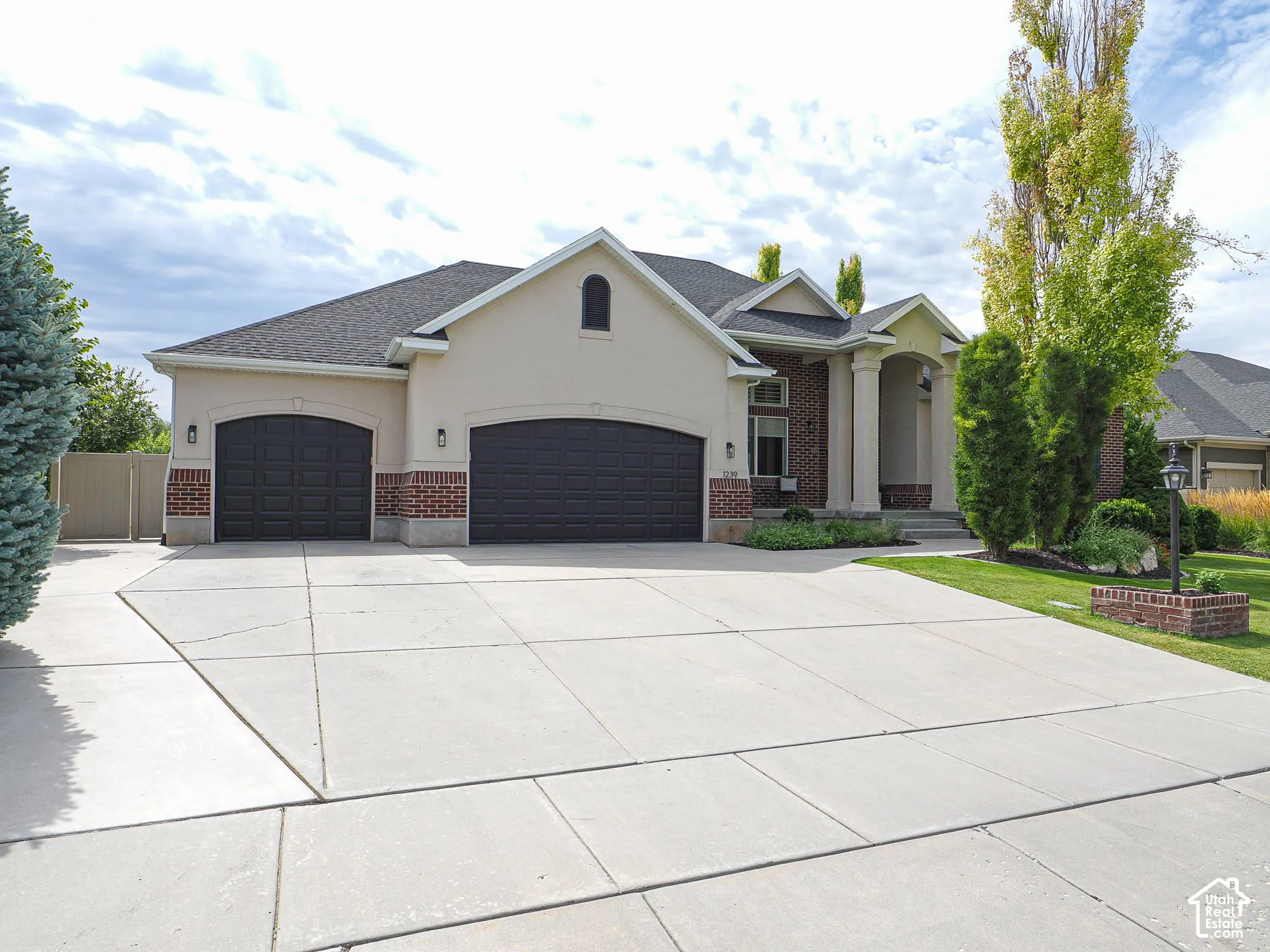 Stucco exterior with brick accents, two-car garage with arched doors, covered entry with columns, concrete driveway, and established landscaping