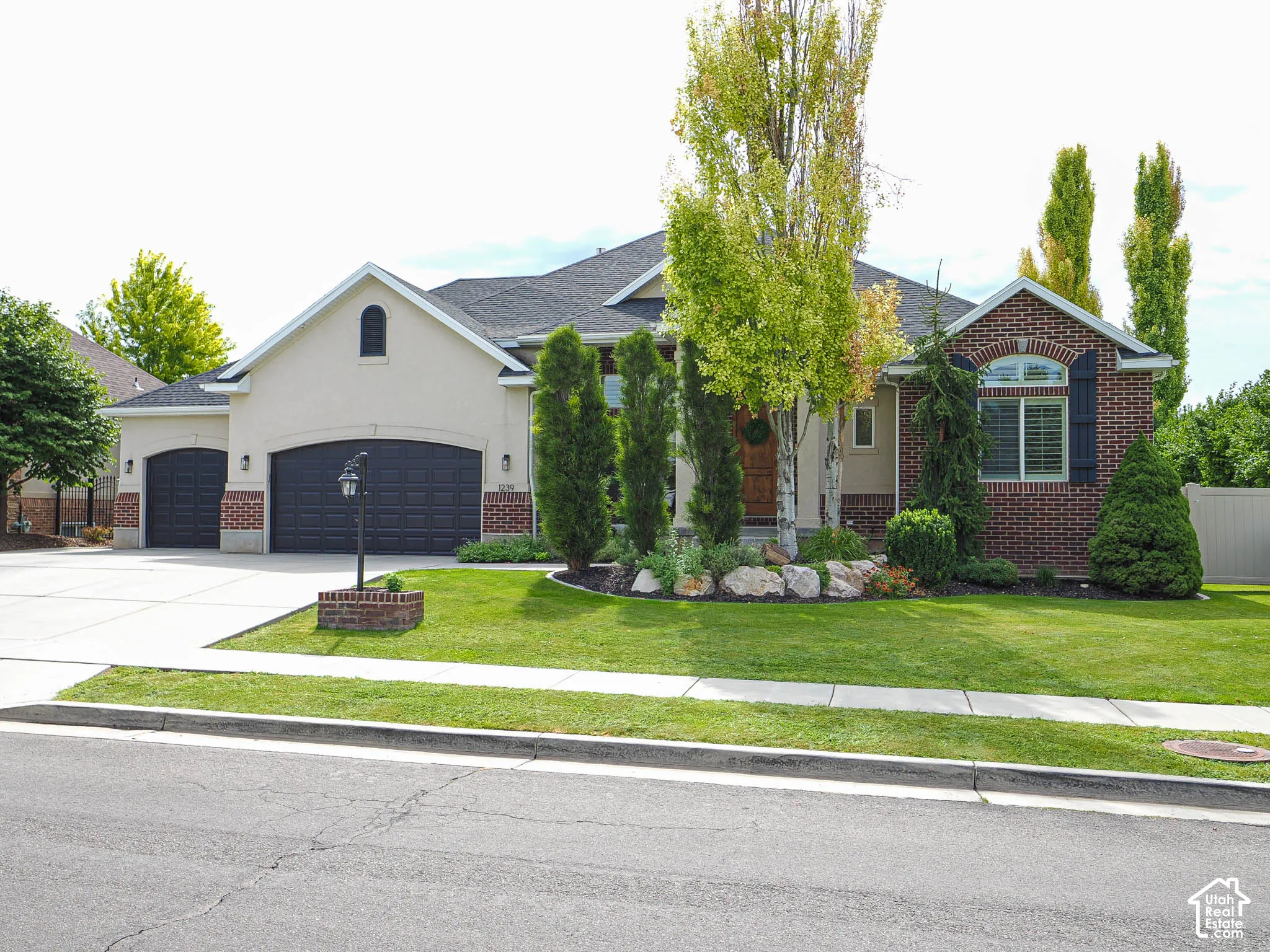 Stucco and brick exterior with arched windows, a two-car garage, concrete driveway, and front lawn landscaping with mature trees