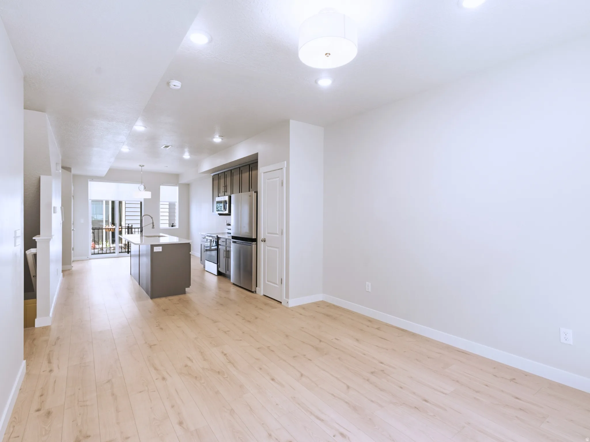 Light wood-finish flooring throughout, recessed ceiling lighting, kitchen island with a rectangular sink and gooseneck faucet, stainless steel appliances, and dark wood-finish cabinetry