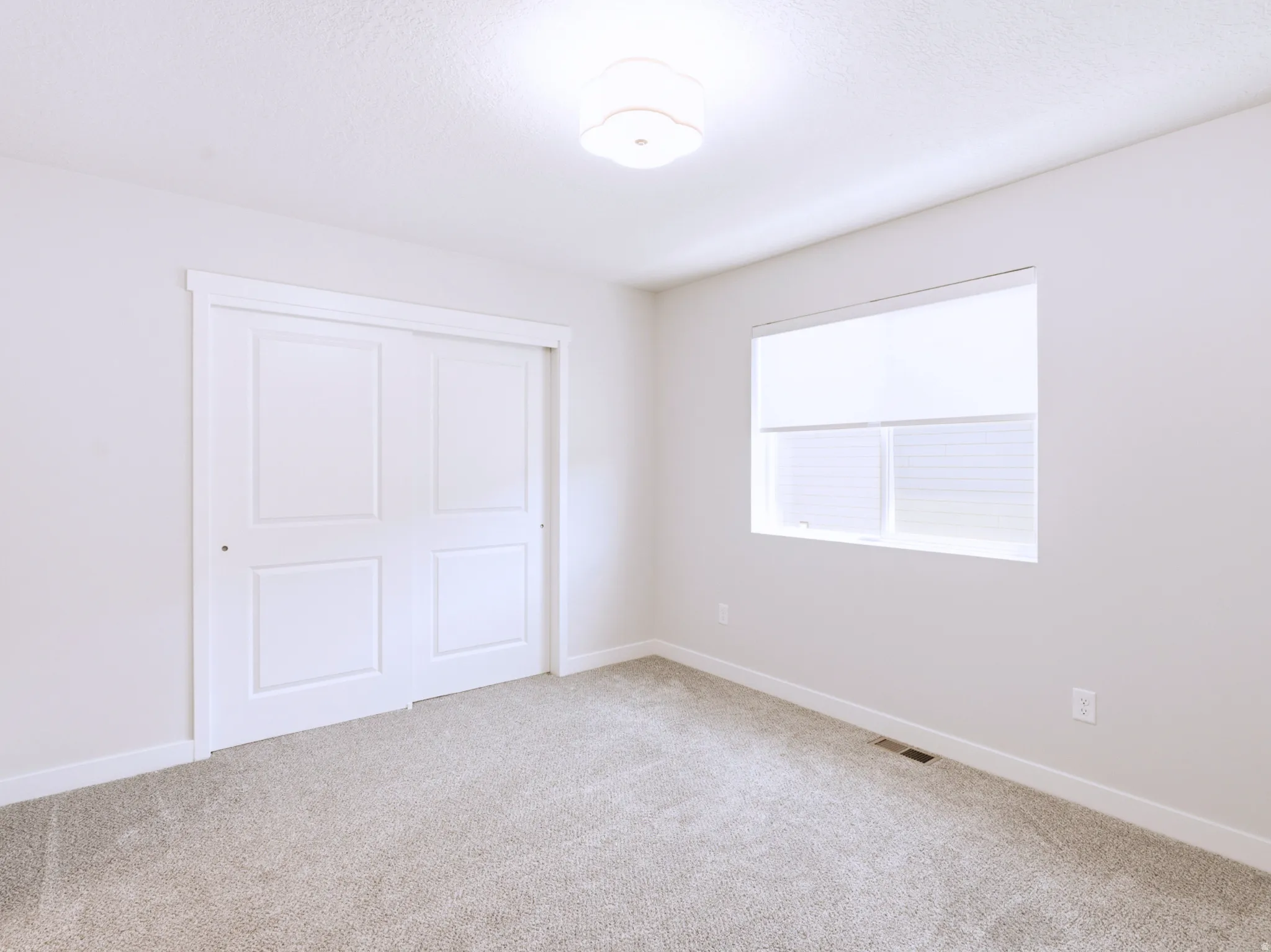 Bright room featuring carpet flooring, a window with roller shade and horizontal blinds, a ceiling-mounted light fixture, and a sliding closet door