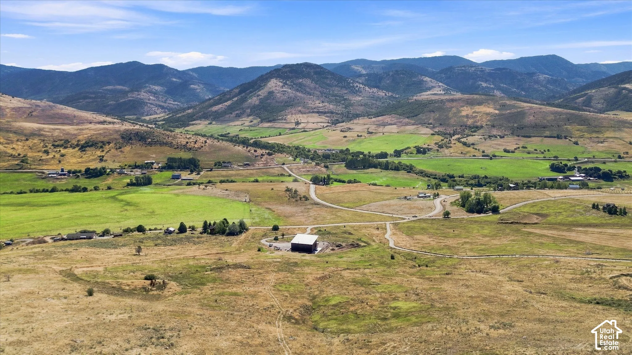 Expansive rural landscape featuring a barn structure, unpaved roads, and distant mountainous terrain