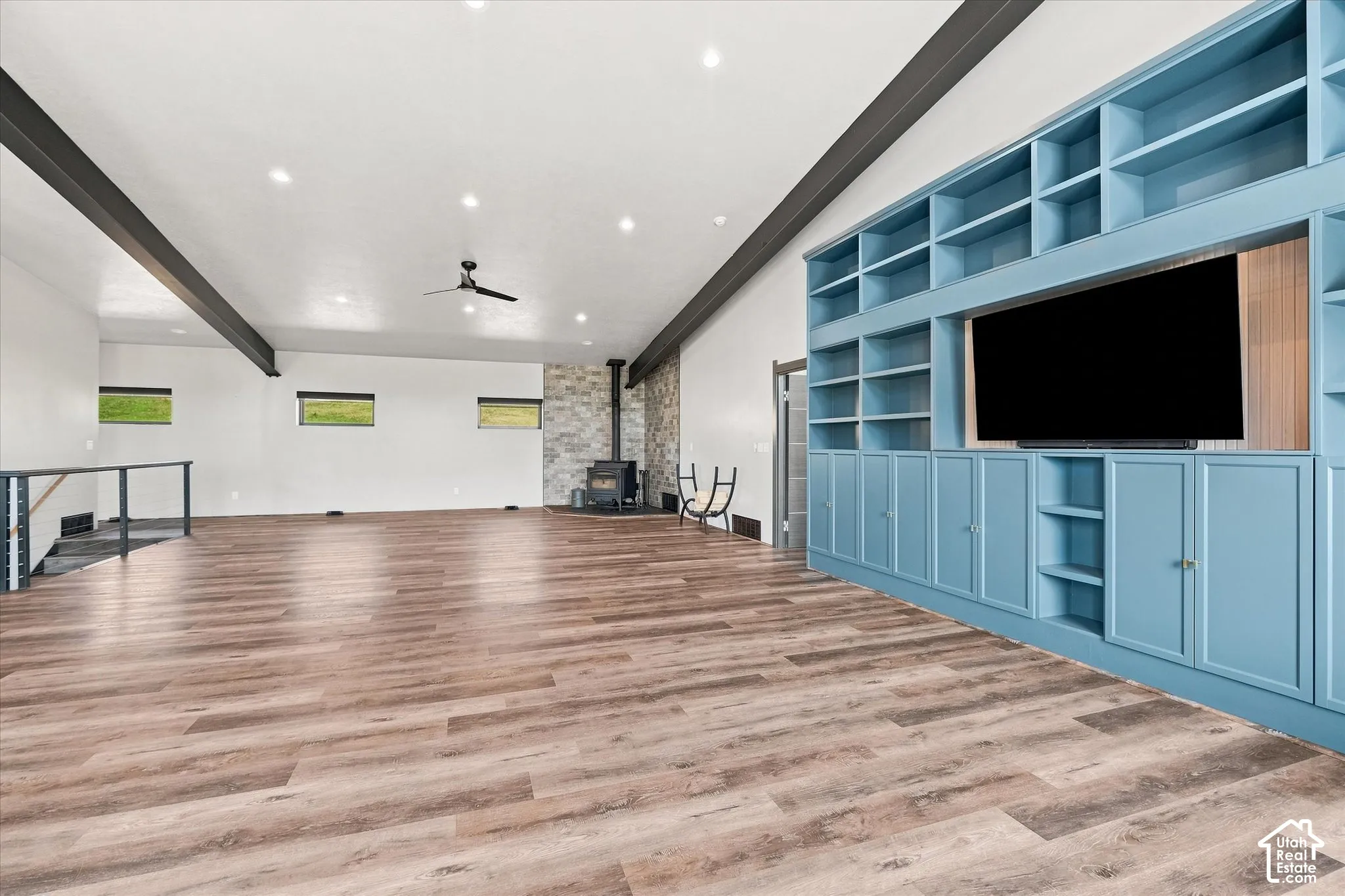 Spacious room featuring wood-finish flooring, a built-in entertainment center with extensive shelving, a wood-burning stove with a stone surround, and exposed ceiling beams