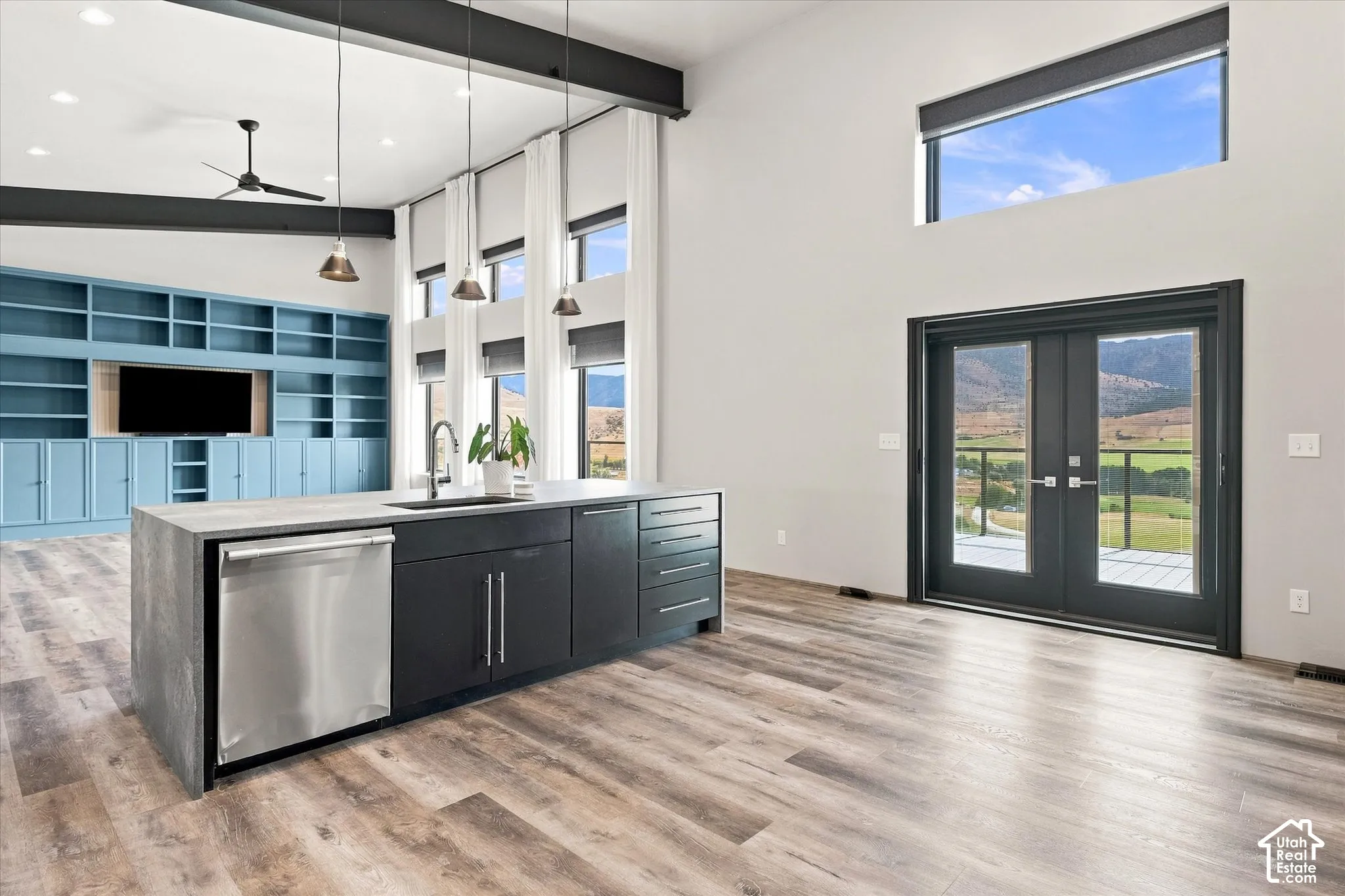 Modern kitchen and great room featuring stainless steel appliances, black cabinetry with brushed metal hardware, a waterfall-edge island with pendant lighting, wood-finish flooring throughout, and expansive glass doors opening to scenic landscape views