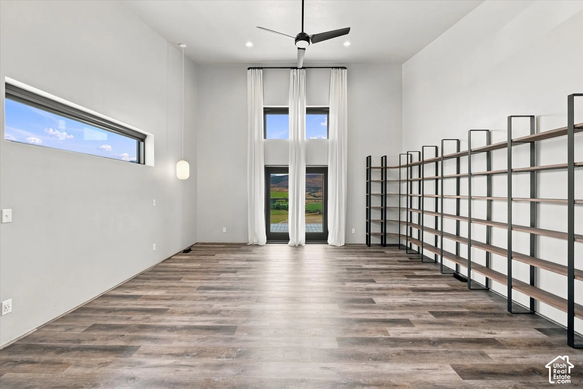 Wood-finish LVP flooring, high ceilings with recessed lighting, black ceiling fan, full-height windows with vertical drapes, and an extensive built-in shelving system