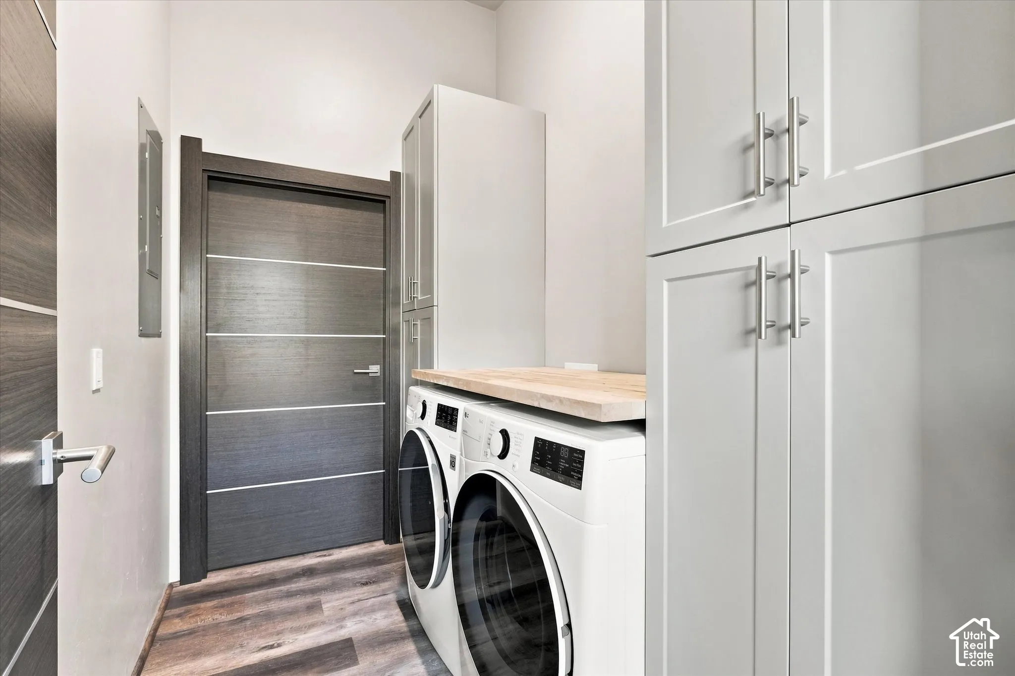 Modern laundry room featuring stacked front-load washers with wood-finish countertop, white cabinetry with chrome hardware, and wood-finish flooring throughout