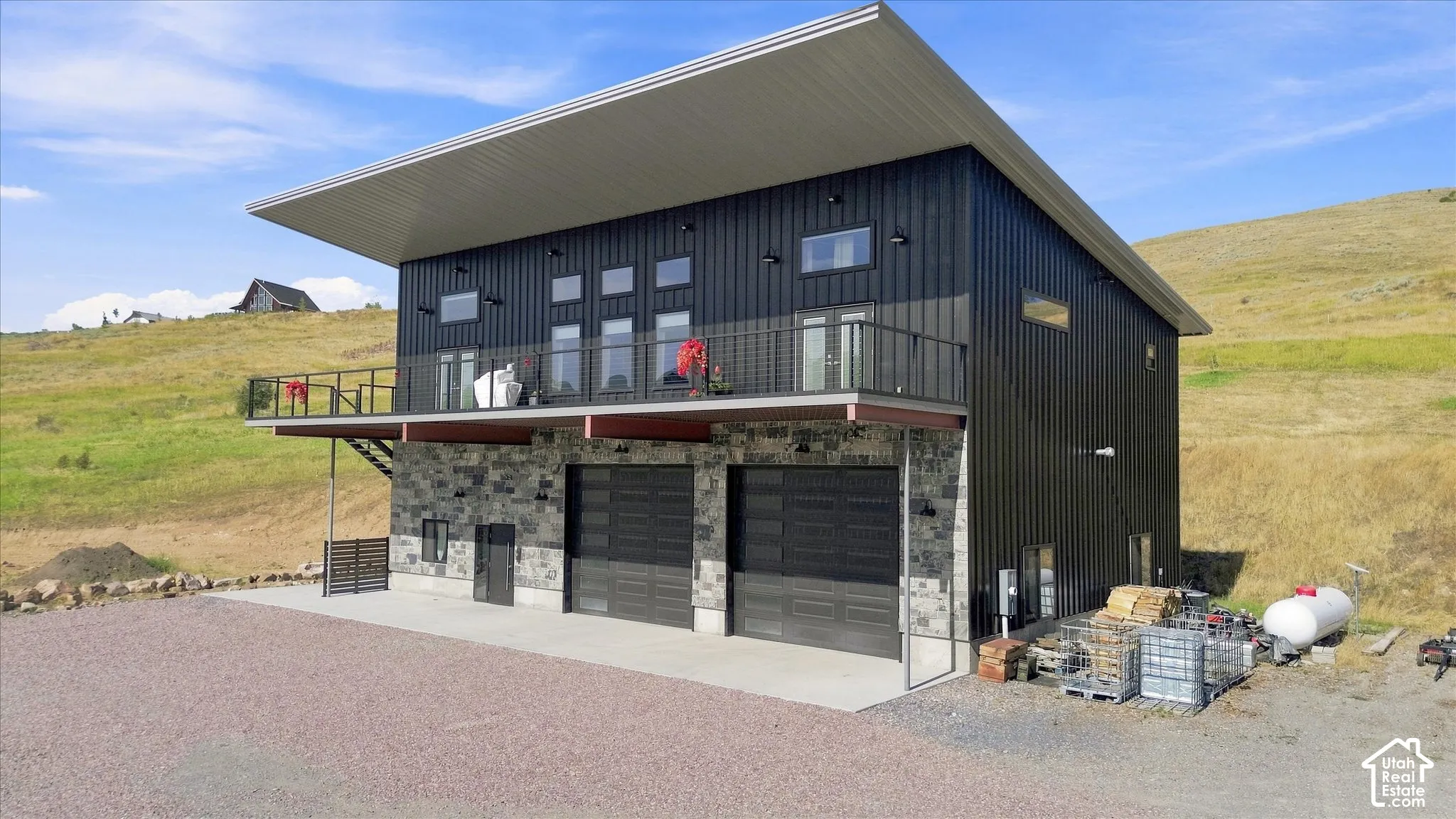 Modern exterior featuring dark vertical siding, stone accents, two large garage doors, an upper-level balcony with metal railing, and an expansive gravel driveway