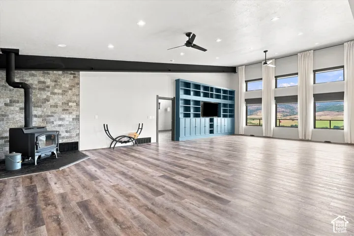 Expansive room featuring wood-finish flooring, a built-in entertainment center, a wood-burning stove with a stone-finish accent wall, and multiple large windows with roller shades