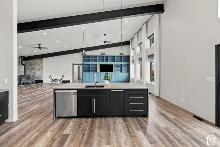 Open-concept kitchen and dining area featuring a large island with black cabinetry and stainless steel appliances, wood-finish flooring throughout, exposed ceiling beams with pendant lighting, and floor-to-ceiling windows with black metal framing overlook