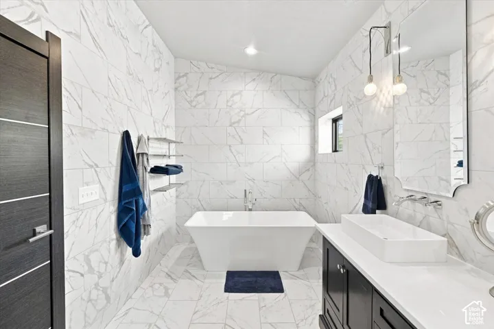 White tile walls and flooring, freestanding soaking tub, dark wood vanity with white countertop, rectangular vessel sink, and wall-mounted faucet