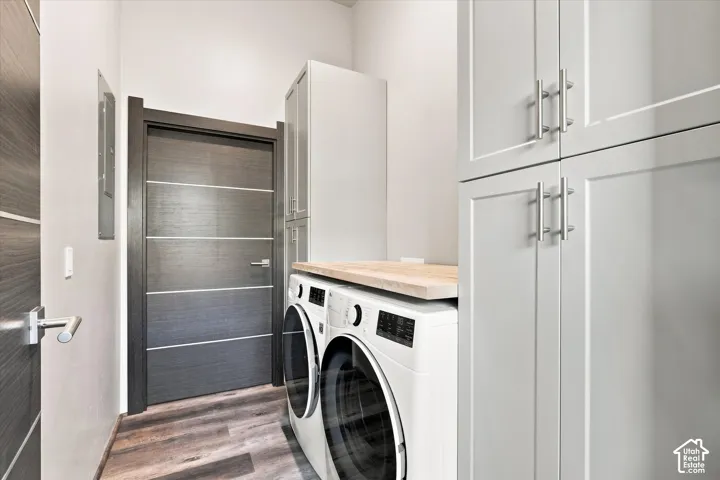 Modern laundry room featuring stacked front-load washers with wood-finish countertop, white cabinetry with chrome hardware, and wood-finish flooring throughout