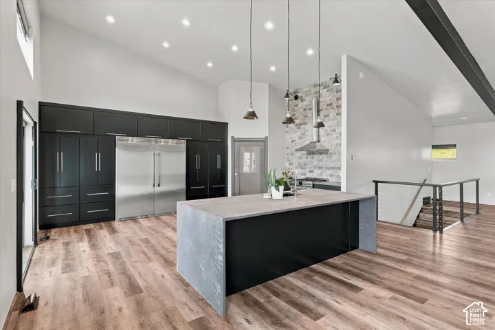 Vaulted ceiling with recessed lighting, wood-finish flooring, a kitchen island with a stone-finish side panel, full-height cabinetry, and a stainless steel refrigerator